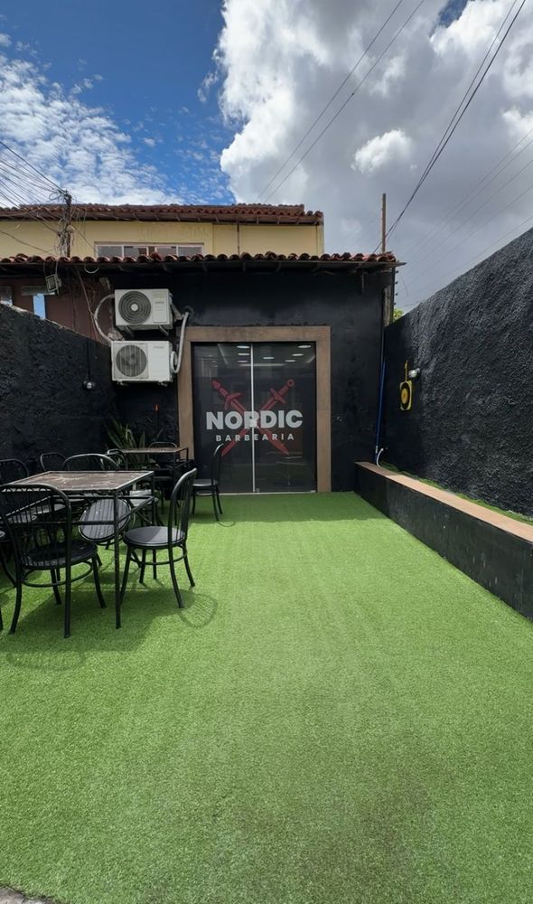 Área externa com gramado, mesas e cadeiras, e a porta de vidro com o logo da Nordic Barbearia
