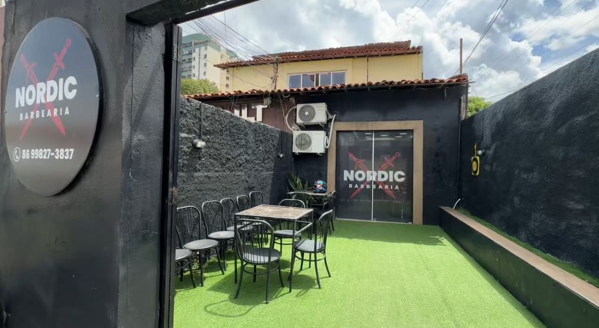 Entrada vista do portão: gramado, mesas e a porta de vidro com o logo da barbearia