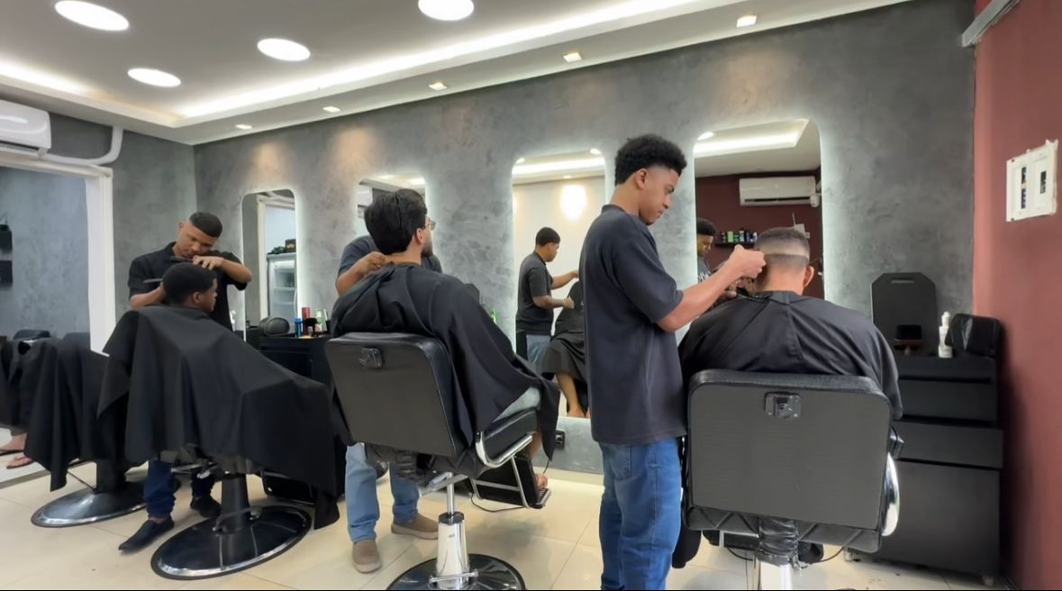 Salão com barbeiros cortando o cabelo de clientes diante dos espelhos iluminados
