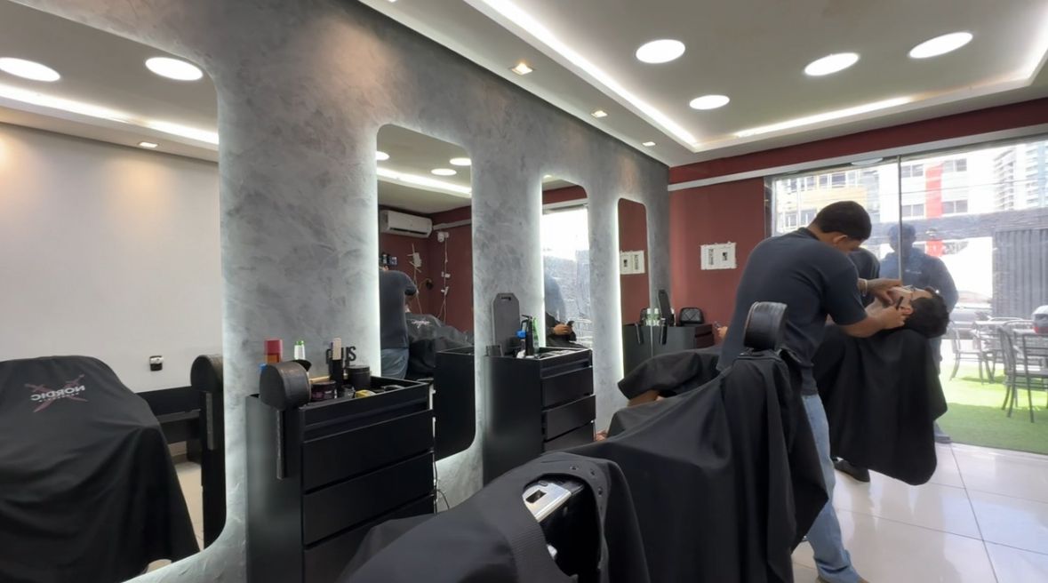Barbeiro fazendo a barba de um cliente, com os espelhos iluminados e a vista para a área externa